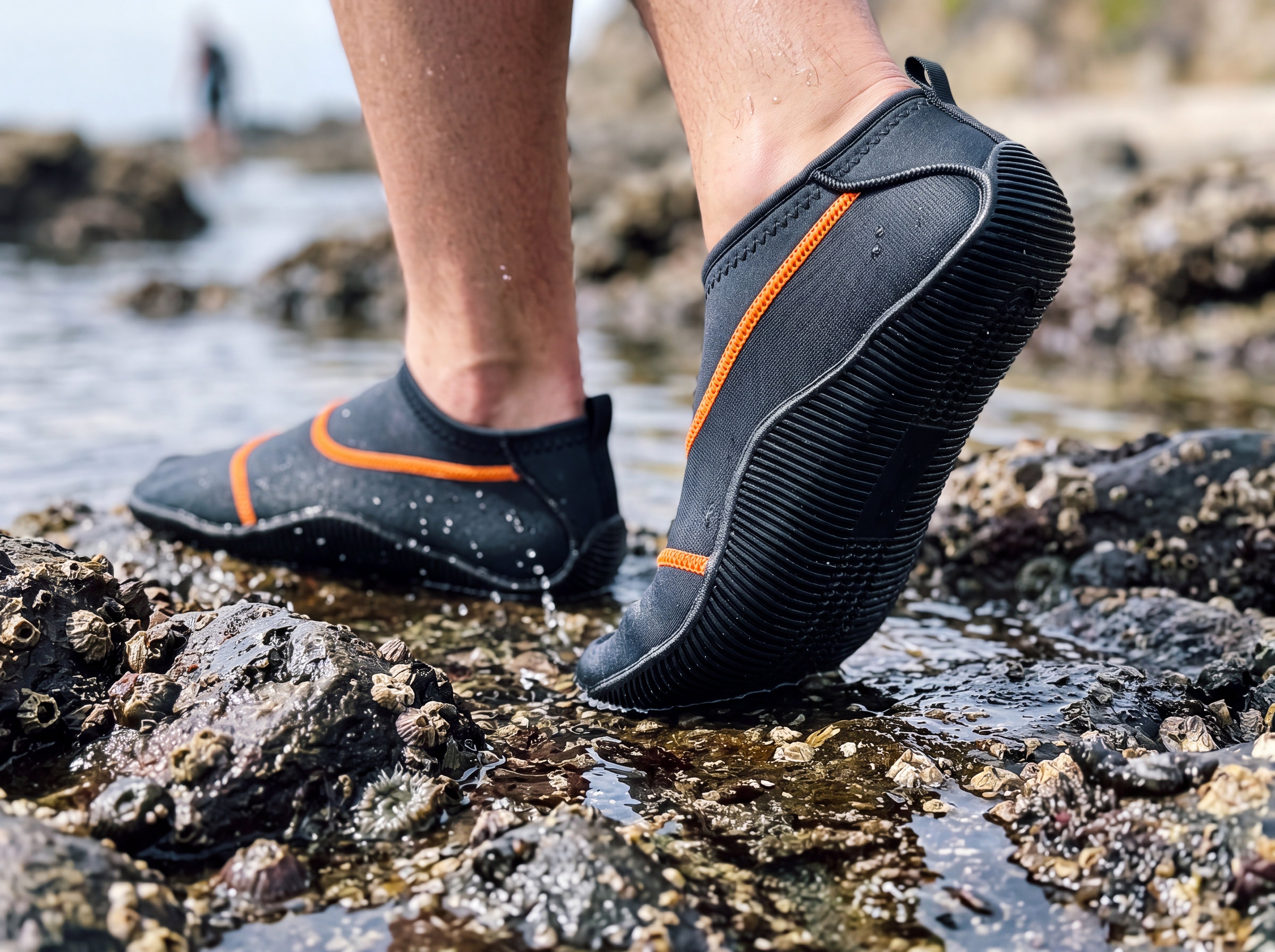 Black Tilos Osmos water socks being worn stepping across wet tidepool rocks, one foot lifted to show the ribbed molded non-slip rubber sole