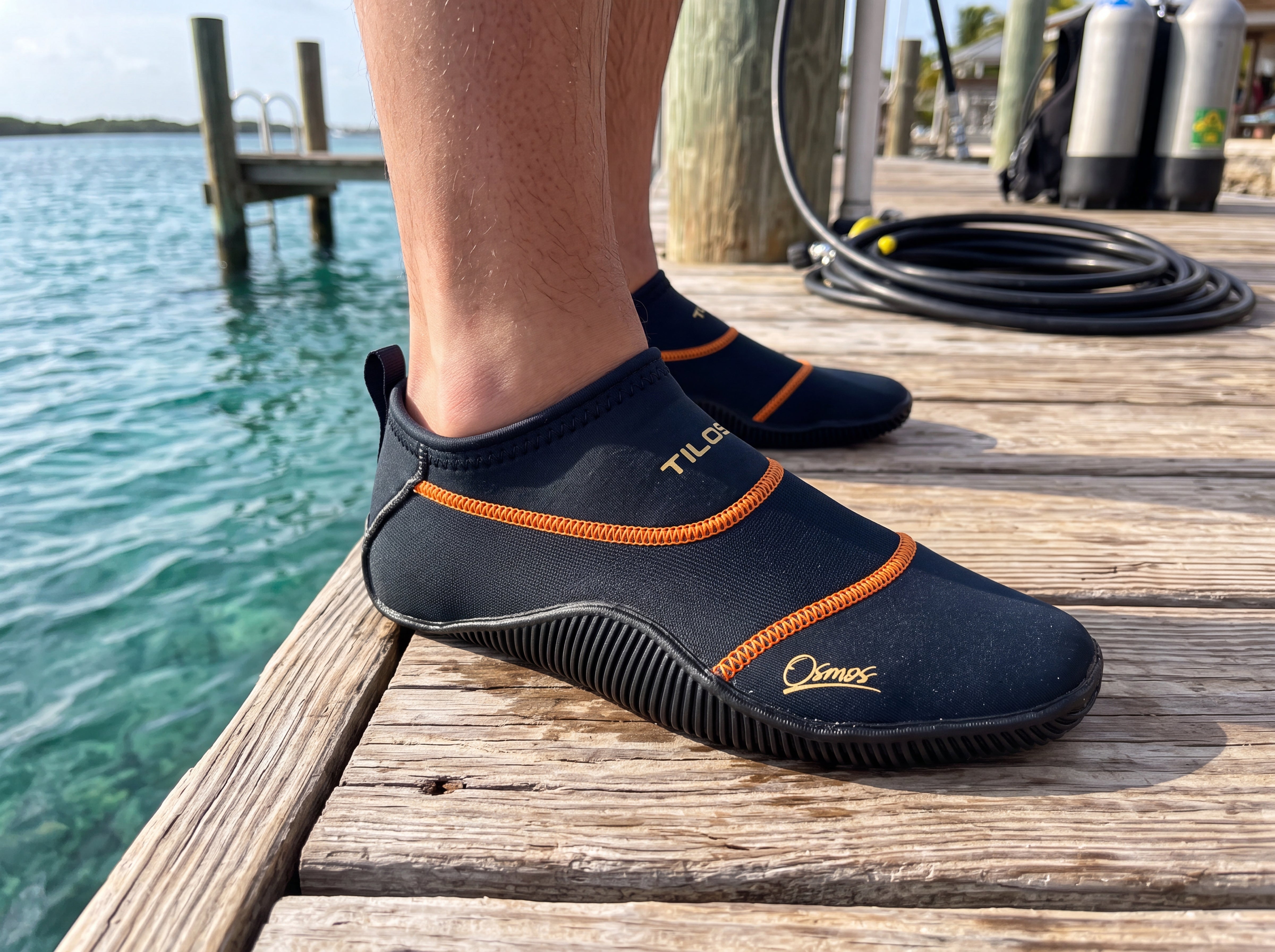 Black Tilos Osmos water socks being worn on a sunlit dive dock, front three quarter view with the gold TILOS wordmark on the ankle collar clearly visible