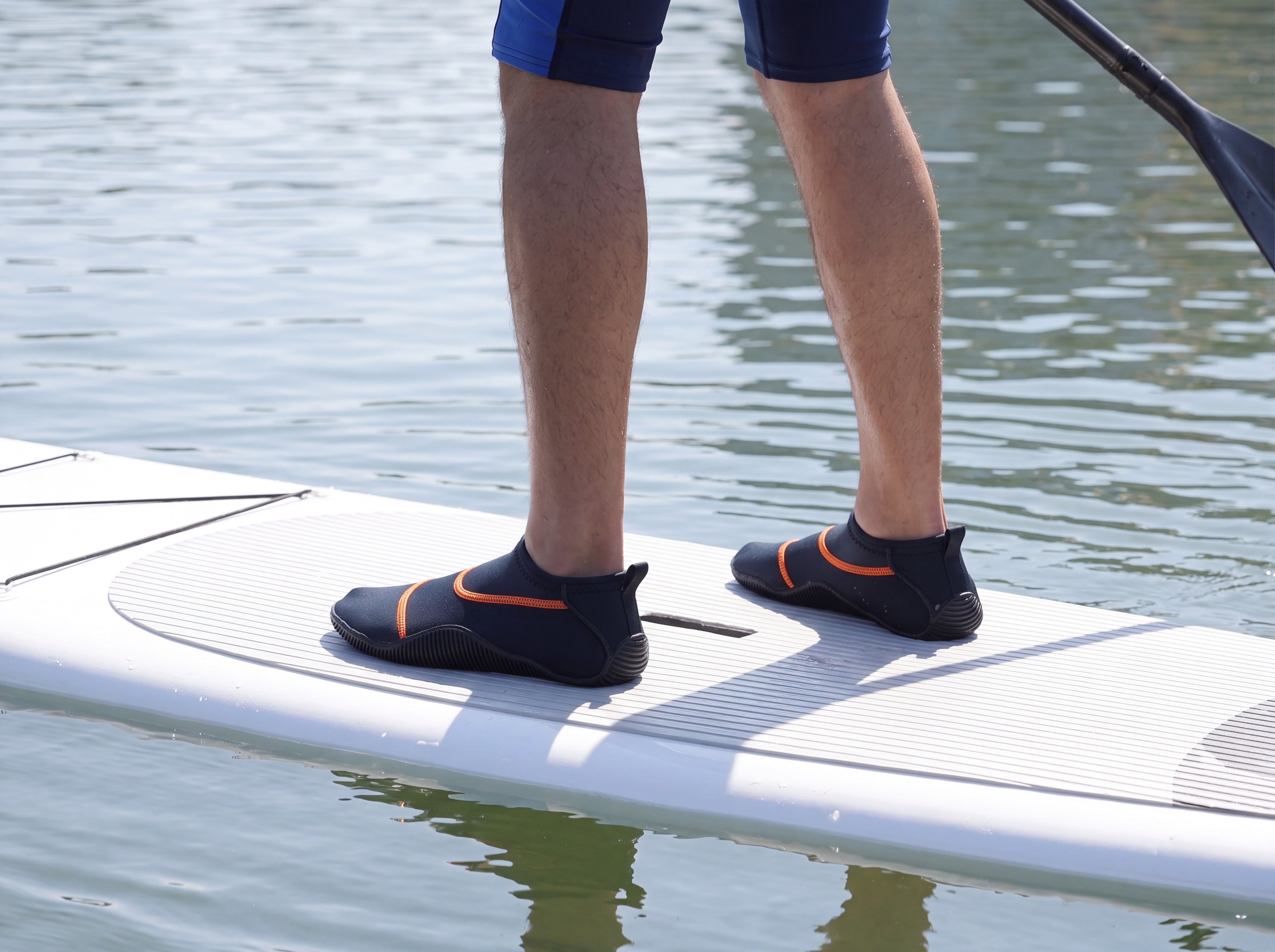 Paddleboarder standing on a stand-up paddleboard wearing black Tilos Osmos non-slip water socks, the ribbed molded soles gripping the deck, seen from behind