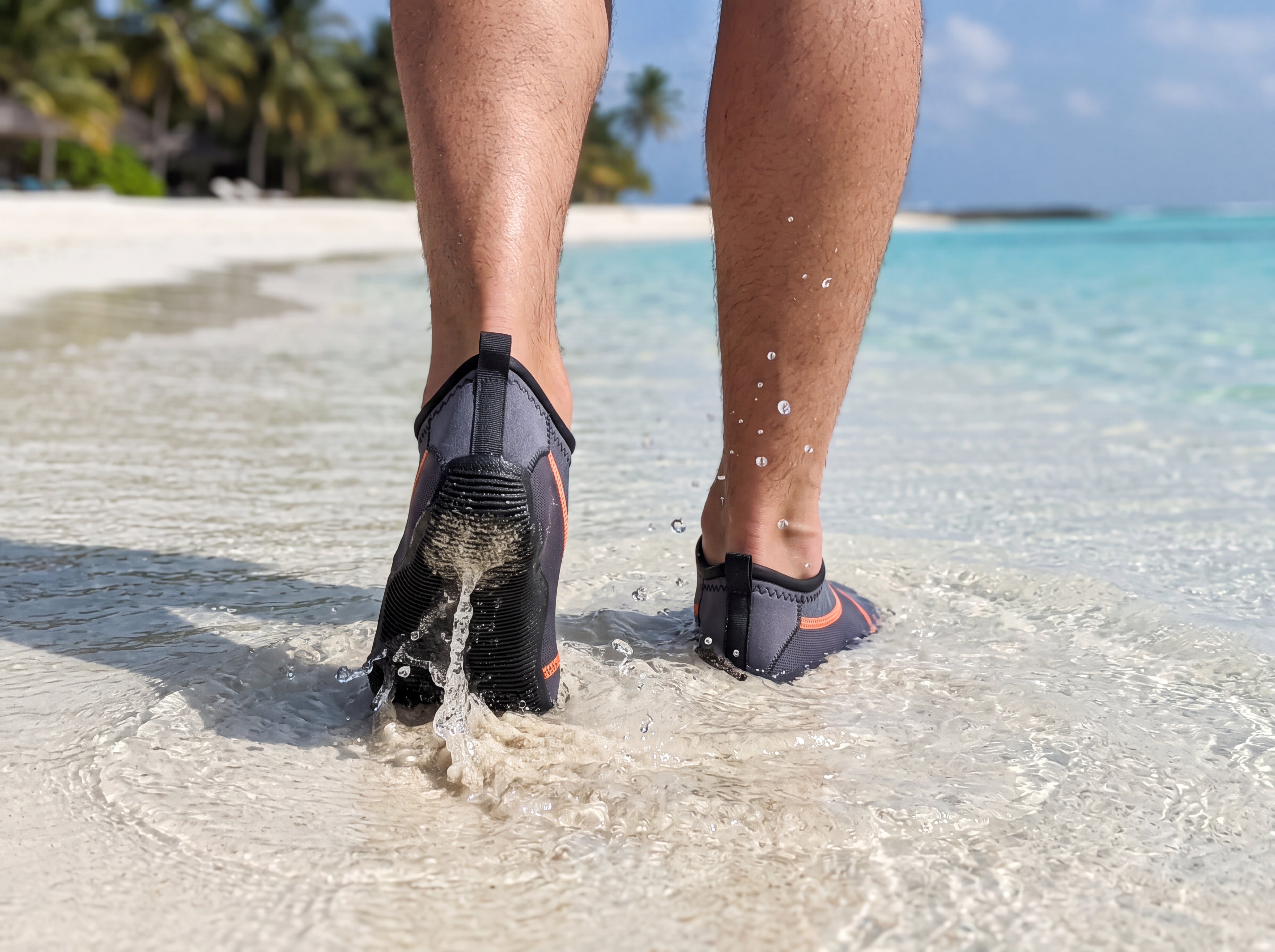 Rear view of a person in the water wading through clear tropical shallows in gray Tilos Osmos water socks, ribbed molded soles and orange piping visible