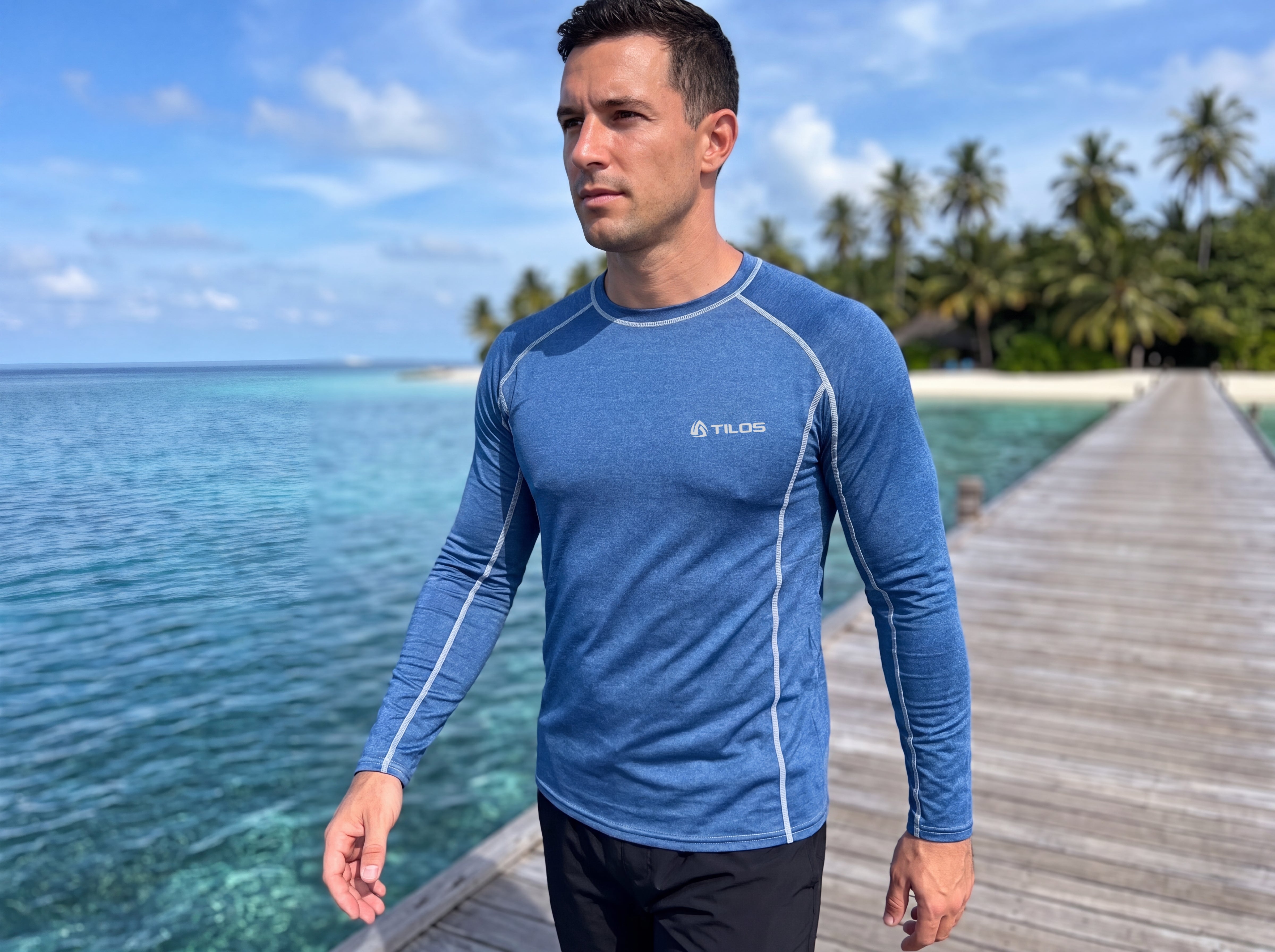 Man walking along a wooden pier over clear water wearing the Tilos Men's 6oz Long Sleeve Rash Guard in Heather Marine blue